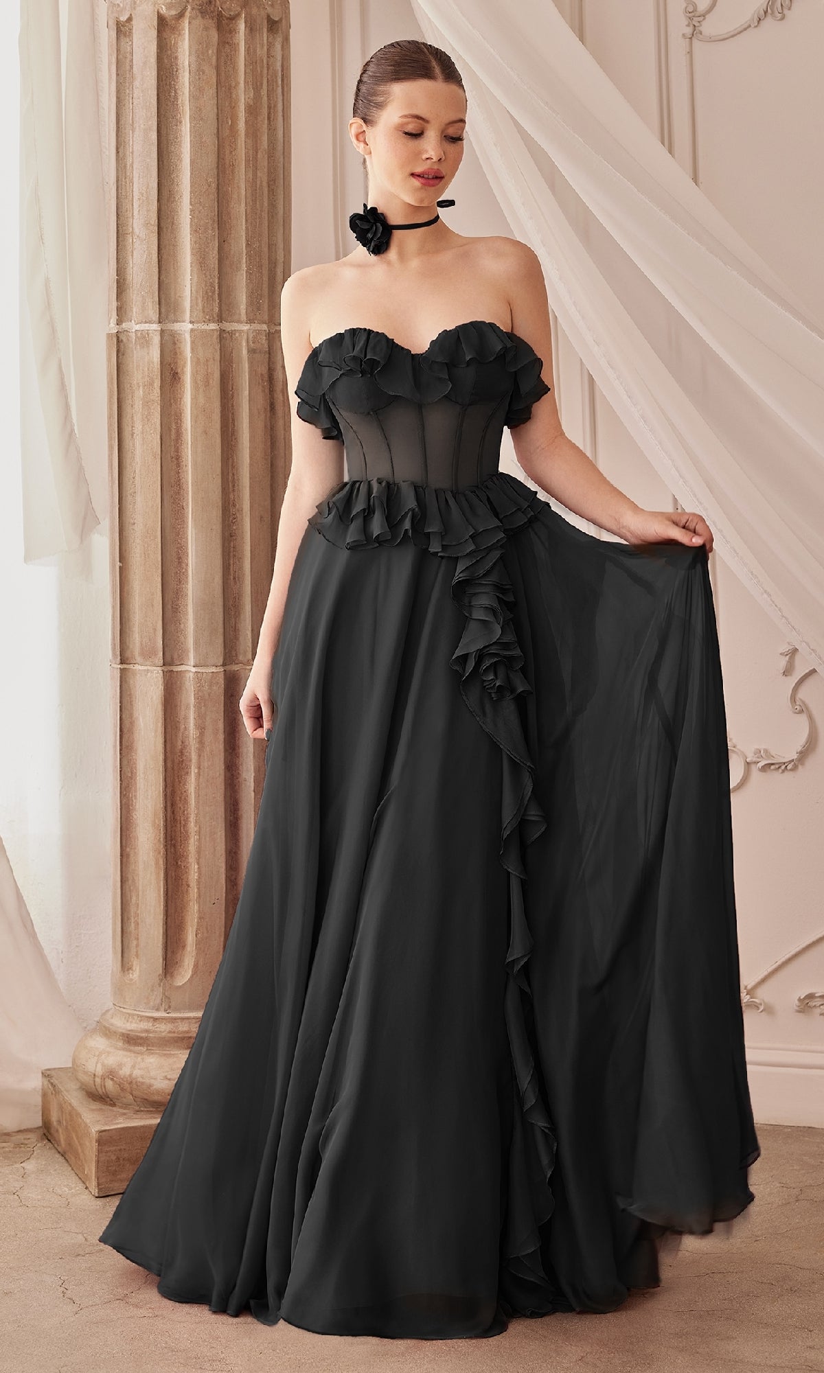A woman stands indoors in the Andrea and Leo A1341 Strapless Ruffled Prom Dress, a black, floor-length gown with a semi-sheer bodice, holding one side of its ruffled skirt.